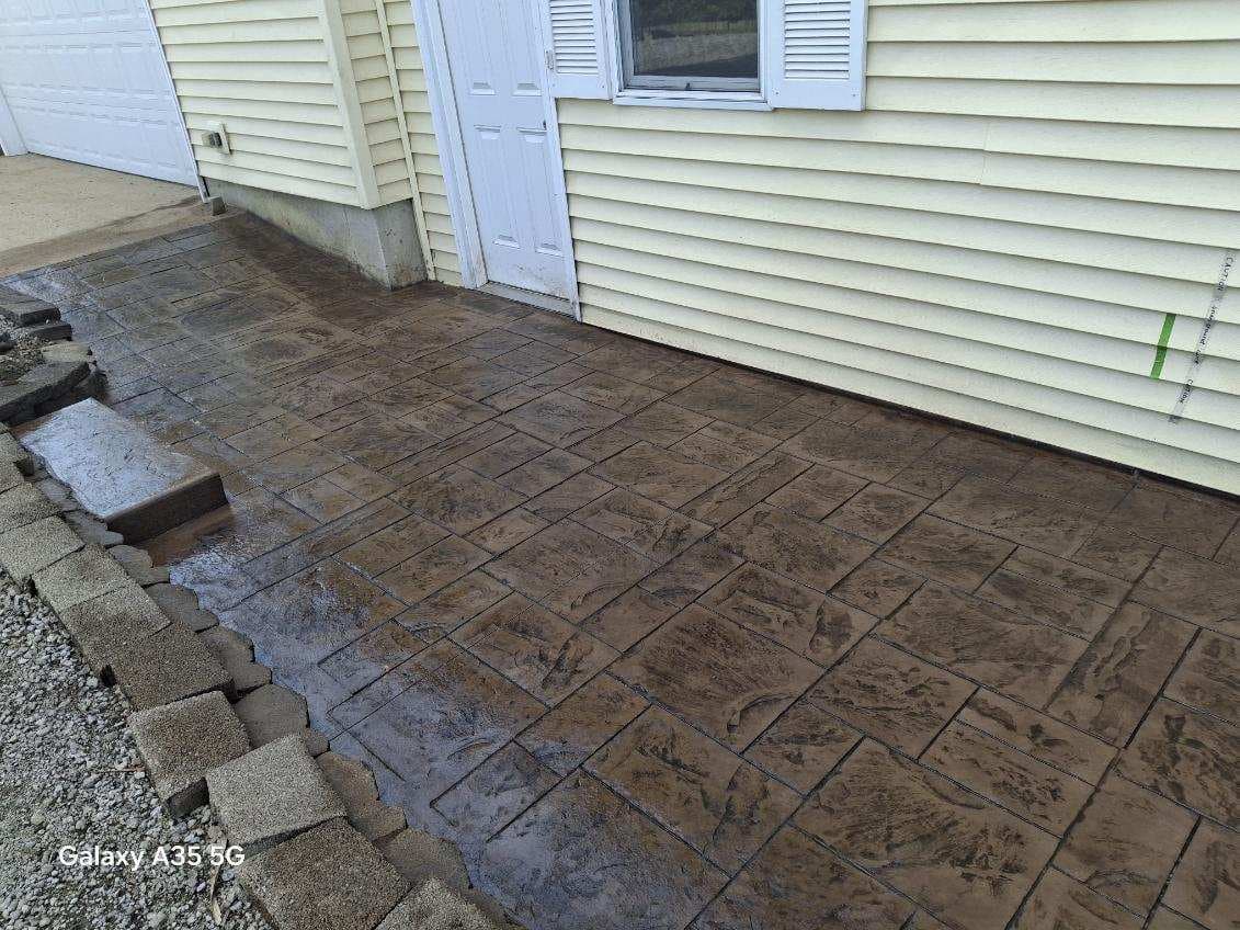 Brown stamped concrete patio with stone border beside a yellow house with white door.